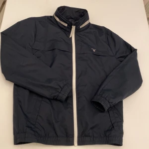 Mörkblå vindjacka från Gant - Snygg mörkblå vindjacka barn från Gant med vit dragkedja och reflekterande detaljer på kragen. Jackan har Gant-logga på bröstet och ett stort tryck på ryggen med 'Sailing Team'. Längd: 60,5cm, ärm 57cm.