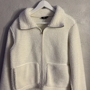 Vit teddyjacka från 157, storlek S - Mysig vit teddyjacka från 157 i storlek S. Jackan har en hög krage, två stora fickor framtill och hel dragkedja. Tillverkad i fluffigt fleece-material som ger en skön och chill vibe. Perfekt för kalla dagar när du vill vara både varm och snygg.