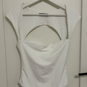 Vit topp med cut out-detalj - Snygg vit topp med unik cut out-detalj framtill och baktill. Toppen har en tight passform och är ärmlös med breda axelband. Perfekt för att skapa en trendig look. Tillverkad i mjukt bomullsmaterial.