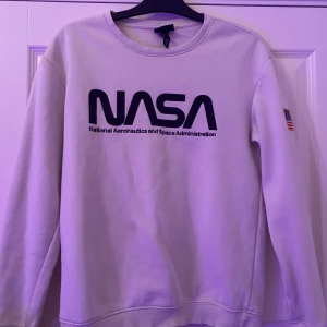 Vit NASA sweatshirt från H&M - Vit sweatshirt från H&M med stor svart NASA-logga på bröstet och amerikansk flagga på ärmen. Tröjan har rund hals, lång ärm och regular fit. Perfekt för dig som gillar rymd och streetstyle.
