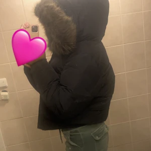 Svart Superdry Everest bomberjacka - Svart bomberjacka från Superdry, modell Everest, med stor huva och fluffig fuskpäls. Jackan har Superdry-logga på ärmen och insidan, samt coola detaljer i guld. Croppad passform och rejäla muddar. Perfekt för kalla dagar och riktigt snygg stil. PRIS KAN DISKUTERAS.