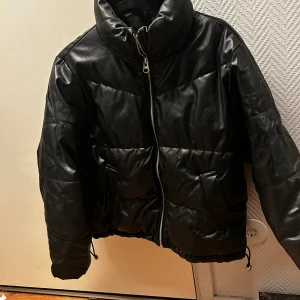 Svart pufferjacka i skinnimitation - Säljer en svart pufferjacka med hög krage och dragkedja framtill. Jackan har två fickor med lock och snörning nedtill för att justera passformen. Materialet är skinnimitation som ger en cool och edgy vibe. Perfekt för kalla dagar när du vill se extra snygg ut.