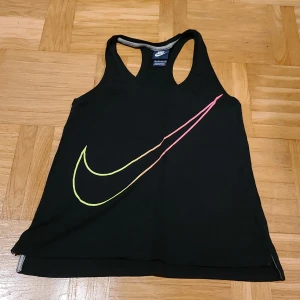 Svart Nike linnetopp XS - Svart linnetopp från Nike i storlek XS med ett stort swoosh-tryck i neonfärger på framsidan. Modellen är ärmlös med brottarrygg och tillverkad i mjuk bomull, perfekt för träning eller chill dagar. Snygg och sportig design med cool färgdetalj.