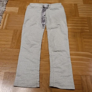 Ljusgrå mjukisbyxor Pink Sweden - Ljusgrå mjukisbyxor från Pink Sweden med satinband som snörning i midjan. Byxorna har rak passform och är tillverkade i mjukt bomullsmaterial. Broderad logga framtill och elastisk midja för skön komfort.