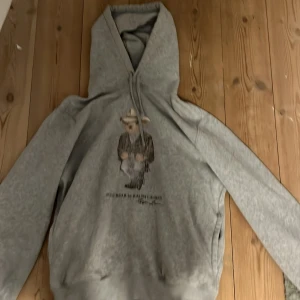 Grå hoodie Polo Bear Ralph Lauren - Grå hoodie från Polo Denim & Supply Ralph Lauren med Polo Bear-tryck på bröstet där björnen har cowboy-outfit. Tröjan har huva med snörning och långa ärmar. Mjuk insida och klassisk passform, perfekt för chill dagar.