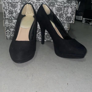 Svarta pumps med hög klack - Snygga svarta pumps med hög klack och plattform. Skorna har en rund tå och är klädda i mockaliknande material som ger en elegant känsla. Perfekta för dig som vill ha en klassisk och stilren look med extra höjd. WIDE FIT pumps med en inlagd comfort sula