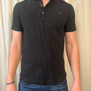 Svart pikétröja Lacoste Sport slim fit - Svart pikétröja från Lacoste Sport i slim fit-modell. Klassisk krage med två knappar och korta ärmar. Diskret Lacoste-logga på bröstet. Tröjan är gjord i mjuk bomull som känns skön mot huden och passar perfekt till jeans eller shorts.