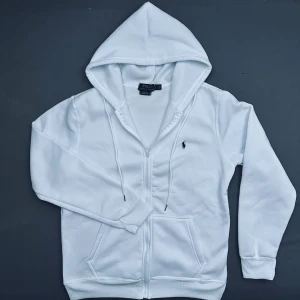 Vit hoodie från Polo Ralph Lauren - Vit hoodie från Polo Ralph Lauren med dragkedja, snörning i huvan och klassisk logga broderad på bröstet. Tröjan har ribbade muddar, två fickor fram och är tillverkad i mjuk bomullsmix. Perfekt för chill dagar och streetwear vibes.