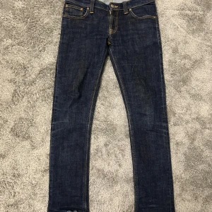 Mörkblå slimfit jeans från Nudie - Snygga mörkblå slimfit jeans från Nudie Jeans med klassisk femficksdesign och kontrastsömmar i orange. Jeansen är tillverkade av 100% ekologisk bomull och har Nudie Jeans logga på bakfickorna. Perfekt för dig som gillar stilrena och smala jeans. Storlek är W28 och L32 men dem är av klippta så det blir typ L30-L31