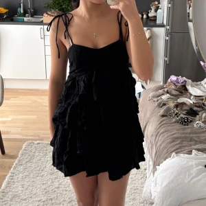 Svart volangklänning med knytband - Supersöt svart kort klänning med volanger och smala axelband som knyts på axlarna. Klänningen har en omlottliknande känsla upptill och flera lager volanger som ger en fluffig och lekfull look. Perfekt för dig som gillar trendiga och feminina plagg.