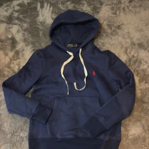 Ralph Lauren hoodie - Snygg marinblå hoodie från Polo Ralph Lauren i slim fit. Tröjan har huva med vita snören, klassisk känguruficka och det röda Polo-logot på bröstet. Tillverkad i mjuk bomull, perfekt för chill dagar. Långa ärmar och ribbade muddar för extra komfort.