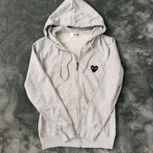 Grå hoodie Comme des Garçons Play - Exklusiv Comme des Garçons Play-hoodie i grått med dragkedja och huva. Ikoniskt svart hjärta fram och stort tryck på ryggen. Storlek S, sitter som XS. Stilren och sällsynt zip – inte många som har denna.