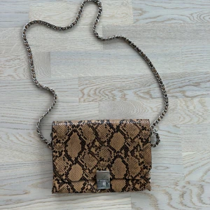 Brun axelväska med ormmönster Zara - Snygg brun axelväska med ormmönster och silverfärgad kedja, från Zara. Väskan har ett stilrent metallspänne framtill och flera fack inuti för smart förvaring. Perfekt storlek för det viktigaste och ger en cool touch till din outfit.