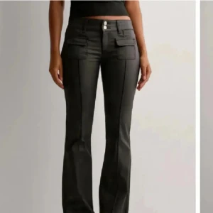 Svarta lowwaist bootcut skinn byxor från Nelly - Använda sparsamt! Inga defekter vad jag ser! Passar runt 166cm långa men såklart kortare än så också! Står ingen storlek men har för mig 32/34 nånstans där, de passar mig som har xs. Snygga svarta bootcut byxor från Nelly med två framfickor med lock och dubbla silverknappar i midjan. Byxorna har låg midja och är tillverkade i ett glansigt syntetmaterial som ger en cool look. Perfekta för dig som gillar trendiga detaljer.