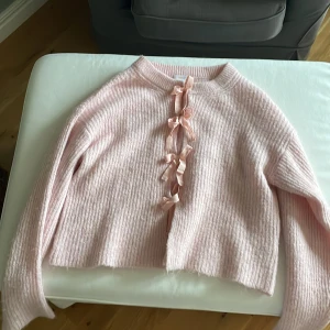 Rosa stickad kofta med rosetter Gina Tricot - Superfin ljusrosa stickad kofta från Gina Tricot med ribbad struktur och tre söta rosetter framtill. Koftan har lång ärm och en croppad passform som ger en trendig look. Perfekt för dig som gillar gulliga detaljer och mjuka material.