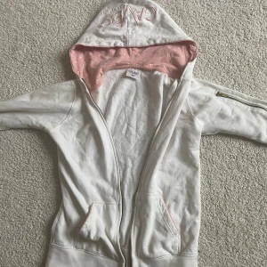 Vit hoodie från Gavelo med rosa detaljer - Snygg vit hoodie från Gavelo i storlek XS med dragkedja och huva. Insidan av huvan är ljusrosa med små hjärtan och Gavelo-loggor. Tröjan har fickor framtill och en dragkedja på ena ärmen. Den har en väldigt 2000s stil/vibe 💘Skriv privat för mått💕dragkedjan är sönder längst ner men är annars i bra skick
