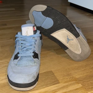 Nike Air Jordan 4 University Blue - Säljer ett par Nike Air Jordan 4 University Blue sneakers i ljusblå mocka med svarta och grå detaljer. Skorna har klassisk snörning, meshpaneler och den ikoniska Jumpman-loggan på tungan och sulan. Perfekta för dig som vill sticka ut med stilrena och sportiga sneakers. Skorna behöver dock tvättas