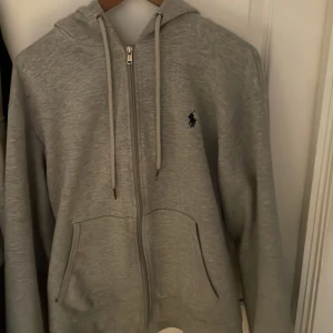 Grå hoodie från Polo Ralph Lauren - Snygg grå hoodie från Polo Ralph Lauren med dragkedja framtill, två fickor och klassisk huva med snörning. Tröjan har lång ärm och den ikoniska Polo-loggan broderad på bröstet. Perfekt för chill dagar och enkel att matcha med jeans eller joggers.