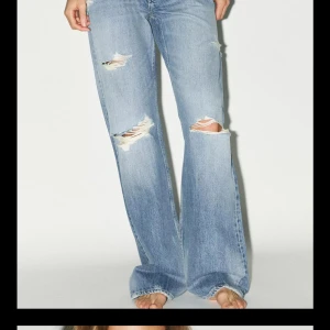 Ljusblå slitna bootcut jeans - Säljer ett par ljusblå bootcut jeans med slitningar och hål på benen. Jeansen har klassisk femficksdesign och är tillverkade i denim. Passformen är normal med hög midja och vida ben. Perfekta för en avslappnad och trendig look. De är avklippta så att de passar någon med 80 i Innerbenslängd och har även en liten slits längst ner