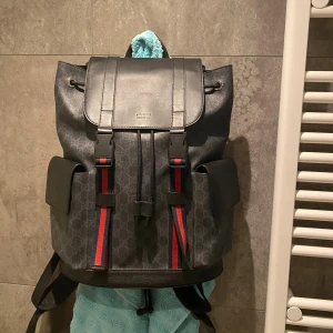 Gucci backpack  Gg supreme - Snygg ryggsäck från Gucci i mörkgrått med klassiskt GG-mönster och svarta detaljer i skinn. Väskan har två sidofickor, lock med spännen och röda/blå ränder framtill. Perfekt för dig som vill ha en lyxig och praktisk väska. Fler bilder kan fås och den har inga defekter då jag har tagit hand om det väldigt väl och den har endast hängt på en vägg som prydnad. Hör gärna av dig! Vid lila minsta fundering. Väskan är själv klart äkta! Pris kan diskuteras vid snabb affär👍