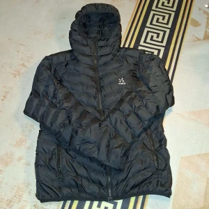 Svart dunjacka från Haglöfs med huva - Svart dunjacka från Haglöfs med snygg quiltad design och praktisk huva. Jackan har hel dragkedja framtill, två sidofickor med dragkedja och diskret Haglöfs-logga på bröstet. Perfekt för kalla dagar och har en clean, sportig look.