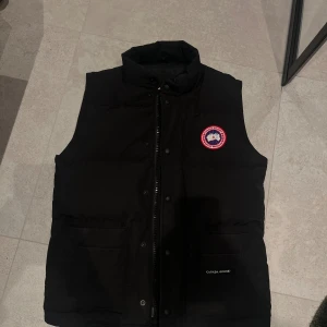 Canada Goose väst - Svart dunväst från Canada Goose med klassisk logopatch på bröstet. Västen har hög krage, dragkedja och tryckknappar framtill samt två stora fickor. Perfekt för lager-på-lager och har en clean, stilren look med diskret branding på fickan.