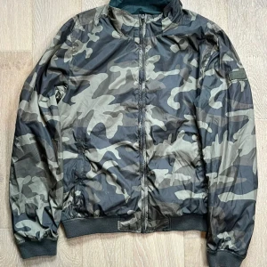 Camouflage vindjacka från WOOLRICH - Snygg vindjacka från WOOLRICH i storlek L med grått och svart camouflage-mönster på ena sidan och helsvart på andra. Jackan är vändbar, har dragkedja framtill och ribbade muddar vid ärmslut och nederkant. Perfekt för dig som gillar streetstyle och vill ha två looks i en jacka.