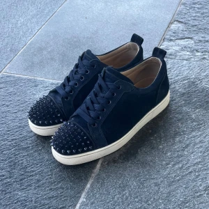 Navy Christian Louboutins  - Säljer ett par svarta sneakers från Christian Louboutin med coola svarta nitar på tån och klassisk röd sula. Skorna har snörning, rund tå och vit platt sula. Ovandelen är i mocka och insidan är beige. Perfekt för dig som vill sticka ut med lyxig streetstyle. Storlek 40 men sitter som 41, ingår: 2 dustbags, nitpåse(utan nitar), skosnörepåse(utan sko snören)