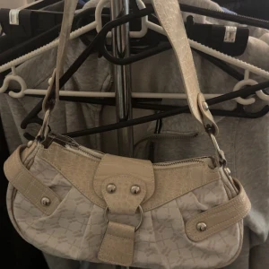 Beige axelväska med krokopräglad detalj - Orginal pris va typ 299, Snygg beige axelväska med krokopräglade detaljer och silverfärgade metalldelar. Väskan har ett subtilt mönster på tyget och dragkedja upptill. Perfekt storlek för det viktigaste och har en cool ringdetalj framtill.