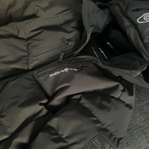Svart Sail Racing dunjacka med huva - Svart dunjacka från Sail Racing med huva och två bröstfickor med dragkedja. Jackan har Sail Racing-logga på bröstet och är tillverkad i syntetmaterial med dunfyllning. Perfekt för kalla dagar och har en clean, sportig look.