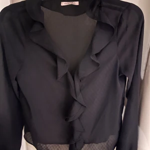 Svart volangblus från Anna Field - Snygg svart blus från Anna Field med volangdetaljer längs v-ringningen. Tunn och luftig känsla, perfekt för dig som gillar en elegant och trendig look. Långärmad modell med feminin touch.