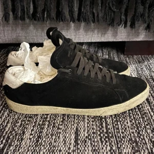 Svarta Saint Laurent sneakers i mocka - Svarta sneakers från Saint Laurent med låg profil och rund tå. Ovandel i mjuk mocka, snörning och vit kontrastdetalj vid hälen. Sulan är beige och har fiskbensmönster undertill. Snygga och stilrena skor som passar perfekt till jeans eller chinos.