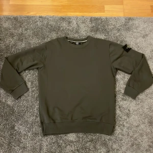 Mörkgrön sweatshirt från Stone Island - Snygg mörkgrön sweatshirt från Stone Island med klassisk rund hals och ribbade muddar. Tröjan har den ikoniska Stone Island-loggan på vänster ärm och är tillverkad i mjuk bomull. Perfekt för dig som gillar stilrena och exklusiva plagg.