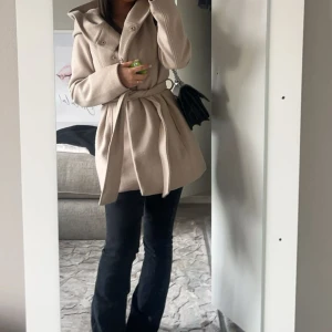 Beige kappa med bälte och huva - Säljer en beige kappa med stor huva och bälte i midjan. Jackan har knappar framtill och är långärmad med en snygg, jättefin passform. Perfekt för dig som gillar stilrena och enkla plagg med en modern touch. Den kommer tyvärr inte till användning har använt 2 gånger 