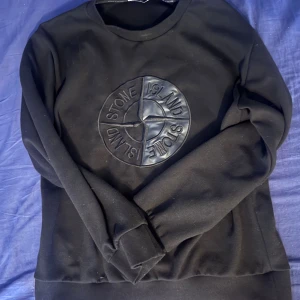 Svart sweatshirt från Stone Island - Svart sweatshirt från Stone Island med stort, upphöjt logomärke i svart på bröstet. Tröjan har rund hals, långa ärmar och ribbade muddar vid ärmslut och nederkant. Materialet känns mjukt och är troligtvis bomull eller polyester. Perfekt för dig som gillar streetwear.