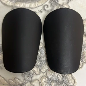 Svarta benskydd för fotboll - Säljer ett par stilrena svarta benskydd som passar perfekt för fotbollsträning och match. De har en slät yta och är formade för att ge bra skydd och komfort. Materialet är hårdplast som tål tuffa tag på planen.