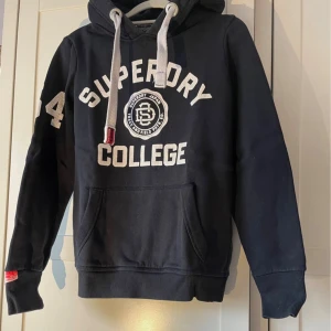 Mörkblå Superdry hoodie(ovanlig) - Blå hoodie från Superdry med stora vita college-tryck framtill och på huvan. Klassisk känguruficka, breda vita snören och ribbade muddar. Perfekt för en sportig och avslappnad stil. Mjuk insida och tydlig logga på ärmen.