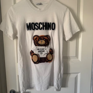 Moschino t-shirt  - Säljer en vit t-shirt från Moschino med broderad nalle på bröstet och texten 'THIS IS NOT A MOSCHINO TOY'. T-shirten har rund hals, korta ärmar och är gjord i mjuk bomull. Perfekt statement-plagg för dig som gillar coola designer och streetstyle.