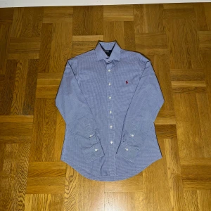 Rutig skjorta från Polo Ralph Lauren - Snygg rutig skjorta från Polo Ralph Lauren i blått och vitt med klassisk krage och knappar framtill. Custom fit-modell med broderad röd logga på bröstet. Långärmad och tillverkad i mjuk bomull, perfekt för dig som gillar stilrena detaljer.