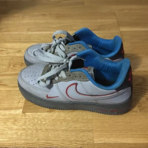 Nike Air Force 1 grå/blå sneakers - Nike Air Force 1 sneakers i grått med blå insida och röda detaljer. Skorna har snörning, perforerad tå och en klassisk röd Swoosh på sidan. Materialet är skinn och syntet, med en platt sula och rund tå. Perfekt för dig som gillar streetwear och sportig stil.