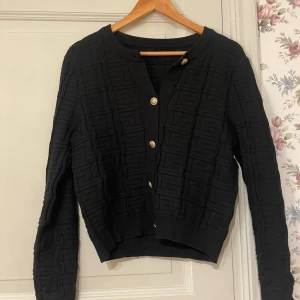 Svart kofta med guldknappar - Stilren svart kofta med markerat mönster och snygga guldknappar framtill. Rund halsringning och ribbade muddar. Fint skick