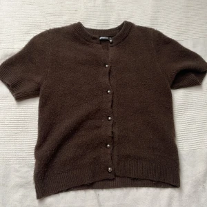 Brun kortärmad kofta från Gina Tricot - Mysig brun kofta från Gina Tricot i storlek XS. Den har korta ärmar, rund hals och pärlknappar framtill. Materialet är stickat och känns mjukt mot huden. Perfekt för lager-på-lager! Använd få gånger 🩷