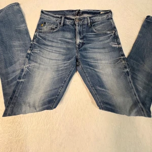 Blå Rifle jeans med tvättad look - Snygga blå jeans från Rifle med tvättad och sliten effekt. Silverfärgad knapp och detaljerad bakficka med Rifle-logga. Jeansen har slim fit , tillverkade i slitstarkt denimtyg.