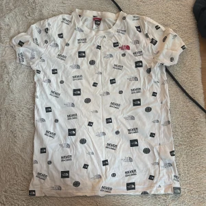 Vit t-shirt The North Face, XL - Snygg vit t-shirt från The North Face i storlek XL Youth/Junior. T-shirten har korta ärmar och är täckt av svart och grått tryck med loggor, texten 'Never Stop Exploring' och globmotiv. Skön och lätt i bomull, perfekt för en avslappnad streetstil.