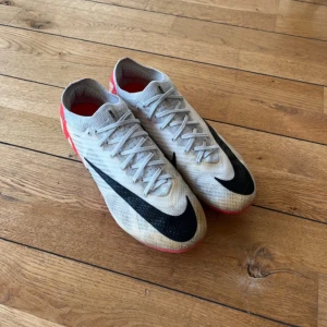 Nike Zoom Mercurial elit fotbollsskor - Nike Zoom Mercurial fotbollsskor i ljusgrått med svarta swoosh-detaljer och röd insida. Skorna har snörning och är tillverkade i syntetmaterial för en lätt känsla på planen. Perfekta för dig som vill ha snabbhet och stil på fotbollsplanen.