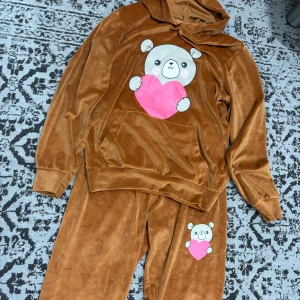 Bruna mjukisbyxor med nalletryck - Bruna mjukisbyxor i mjukt sammetstyg med ett gulligt nalletryck som håller ett rosa hjärta på ena benet. Byxorna har en loose passform och är perfekta för chill dagar hemma. Matchar med hoodie i samma stil för en söt och mysig look.