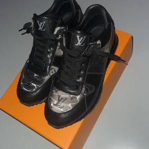 Louis Vuitton skor äkta - Snygga svarta Louis Vuitton sneakers med klassiskt monogrammönster i grått och silverdetaljer. Skorna har rund tå, snörning och är tillverkade i skinn med LV-logga på tungan och hälen. Perfekta för dig som vill sticka ut med exklusiv streetstyle.