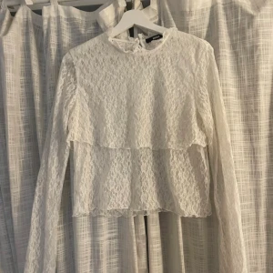 Vit spetsblus med volang - Säljer en vit blus från Bik Bok i genombruten spets med lång ärm och hög hals. Blusen har en söt volangdetalj över bröstet och knäppning bak i nacken. 