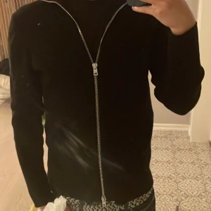 Svart kofta med dragkedja från Ljung - Svart stilren kofta från Ljung by Marcus Larsson i storlek M. Tröjan har en lång silverfärgad dragkedja framtill och är långärmad. Materialet känns mjukt och har en lätt ribbad struktur. Perfekt för dig som gillar minimalistisk och clean look.