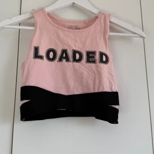 Rosa croppad linnetopp med LOADED-tryck - Säljer en rosa croppad linnetopp med svart LOADED-tryck framtill och bred svart kant nedtill. Toppen är ärmlös och har en sportig vibe, perfekt för dig som gillar att sticka ut. Mjuk bomullskvalitet och rund halsringning.